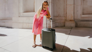 Smiling woman in a pink dress posing with a durable, large suitcase from Samsonite in a sunny outdoor setting.